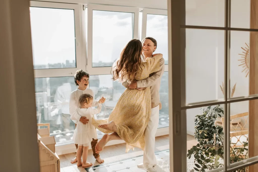 How to Honor Your Mental Health This Thanksgiving-Image of a happy family, well dressed to celebrate Thanksgiving, parents hugging, boy and girl smiling, in an environment that is illuminated by flashes of light through a glass window.